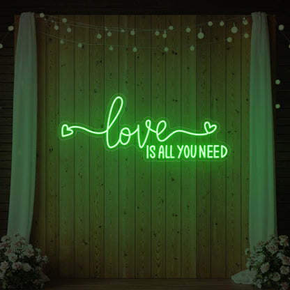 Photo of the 'Love Is All You Need' neon sign glowing in green against a styled interior wall.