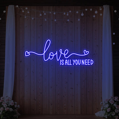 Photo of the 'Love Is All You Need' neon sign glowing in deep blue against a styled interior wall.