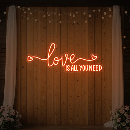 Photo of the 'Love Is All You Need' neon sign glowing in golden yellow against a styled interior wall.