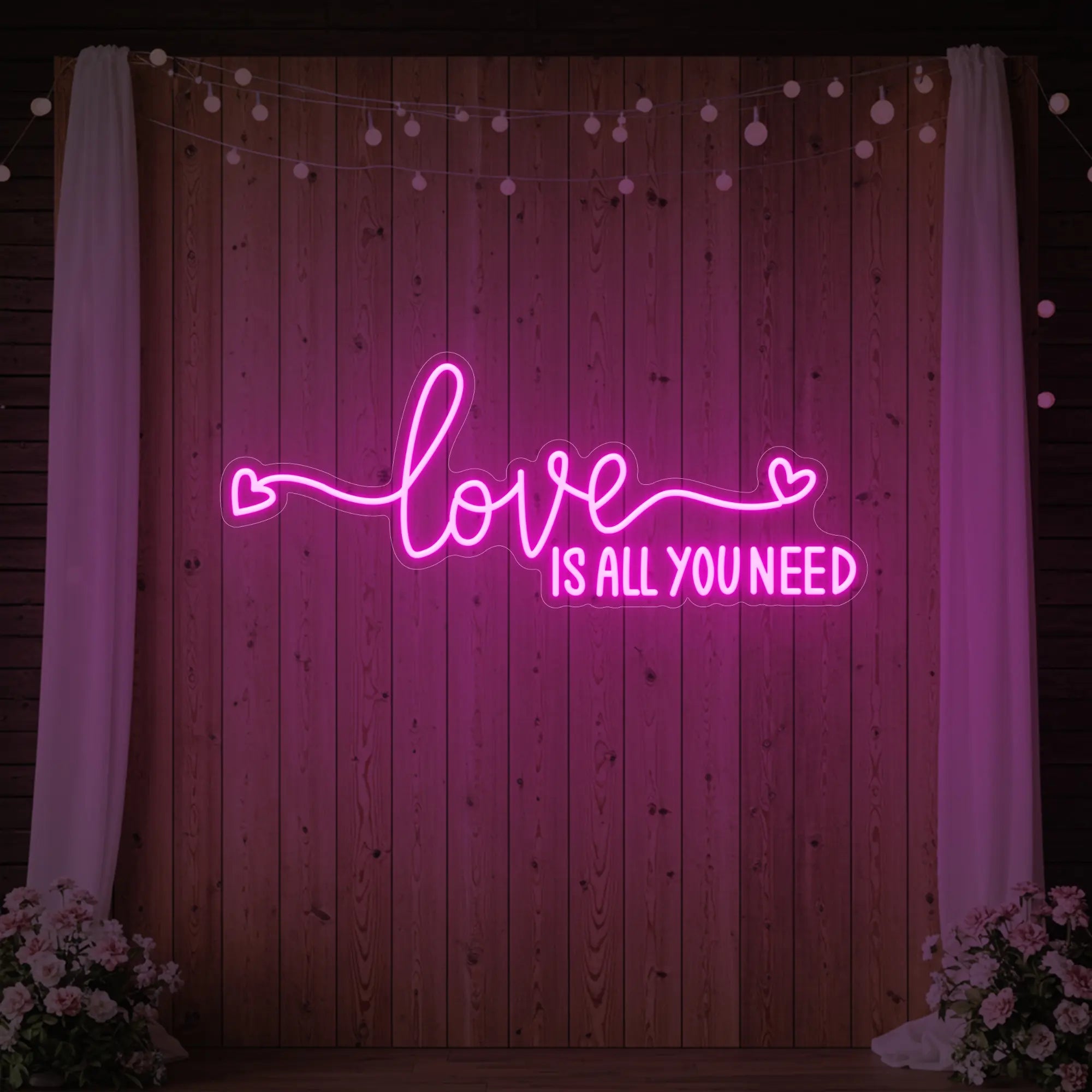 Photo of the 'Love Is All You Need' neon sign glowing in light pink against a styled interior wall.