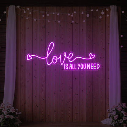 Photo of the 'Love Is All You Need' neon sign glowing in deep pink against a styled interior wall.