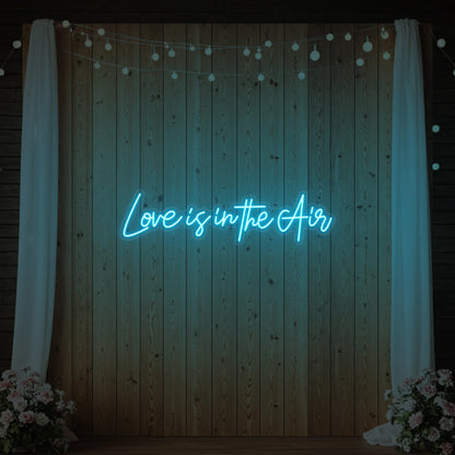 Photo of the 'Love Is In The Air' neon sign glowing in ice blue against a styled interior wall.