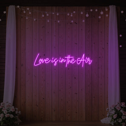 Photo of the 'Love Is In The Air' neon sign glowing in deep pink against a styled interior wall.