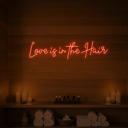 Photo of the 'Love is in the Hair' neon sign glowing in golden yellow against a styled interior wall.
