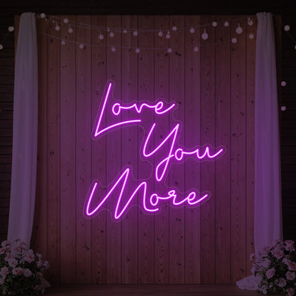 Photo of the 'Love You More' neon sign glowing in deep pink against a styled interior wall.