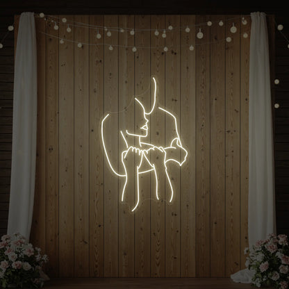 Photo of the 'Lovers Holding Each Other' neon sign glowing in warm white against a styled interior wall.