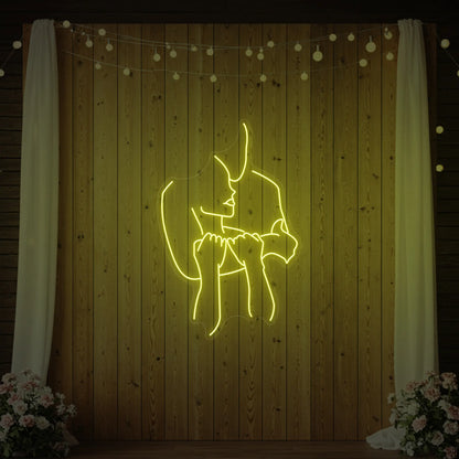 Photo of the 'Lovers Holding Each Other' neon sign glowing in lemon yellow against a styled interior wall.