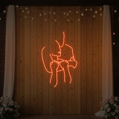Photo of the 'Lovers Holding Each Other' neon sign glowing in golden yellow against a styled interior wall.