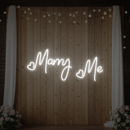 Photo of the 'Marry Me' neon sign glowing in cool white against a styled interior wall.