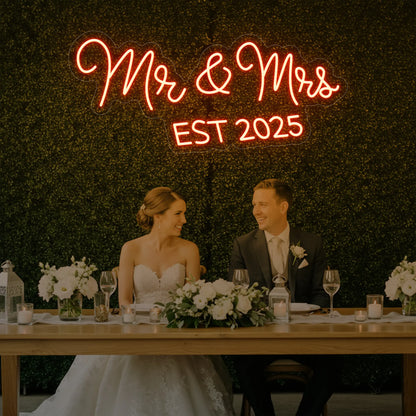 Photo of the 'Mr & Mrs EST' neon sign glowing in golden yellow against a styled interior wall.
