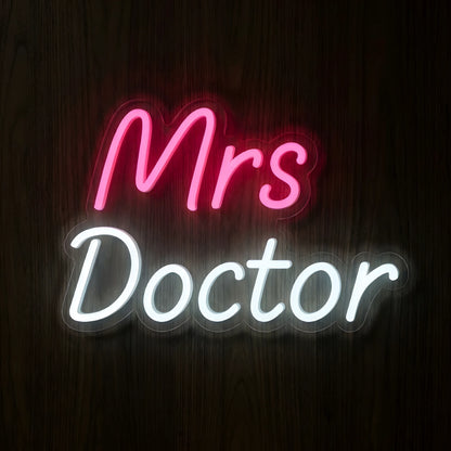 Mrs Doctor Script LED Neon Sign featuring glowing pink Mrs lettering above elegant white Doctor cursive text on clear cut-to-shape acrylic backing against a dark wooden wall.