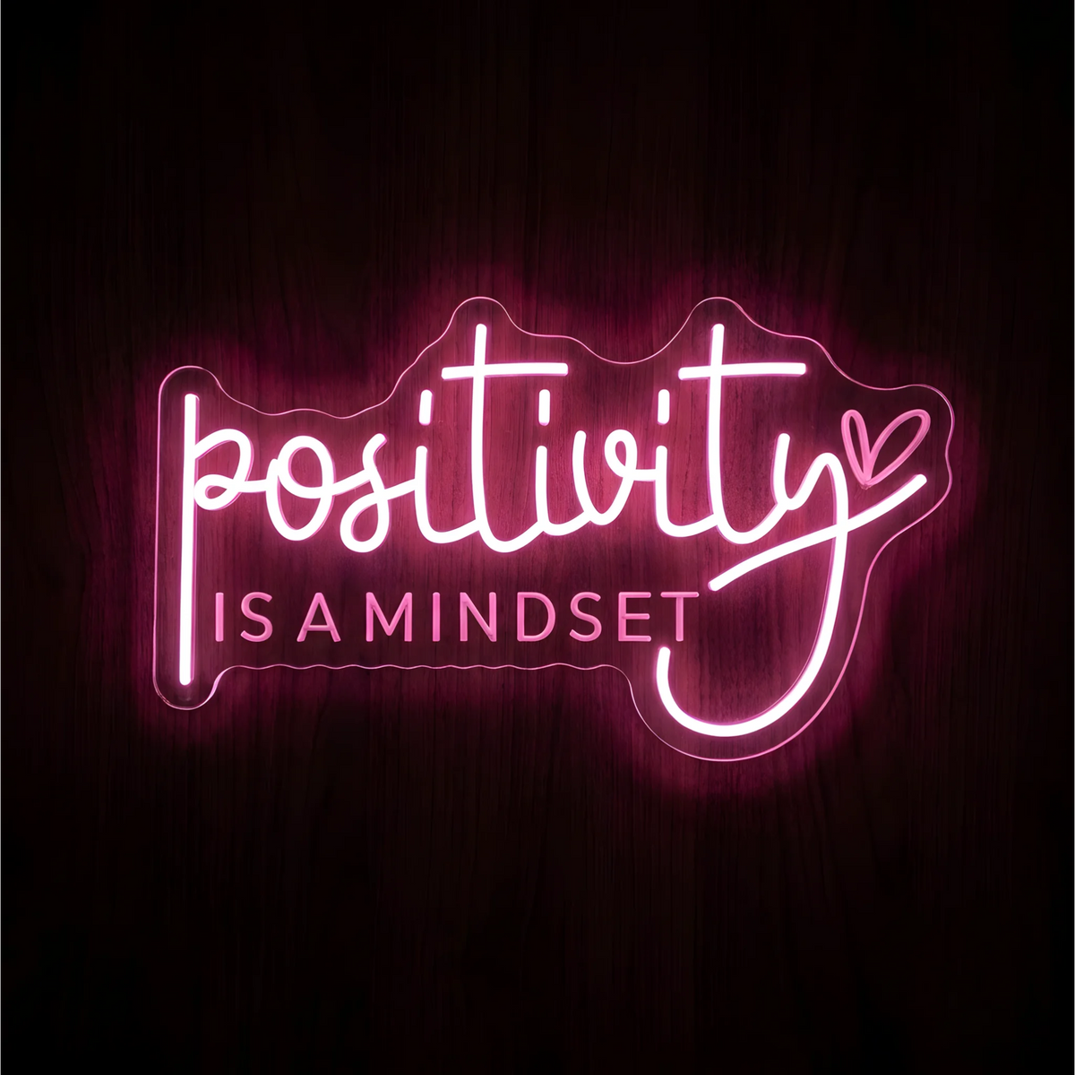 Positivity Is a Mindset LED Neon Sign in pink and white lettering with a small heart at the end of the script, displayed on clear acrylic against a dark wood background, suited to bedrooms and wellness spaces.