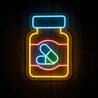 Prescription Pill Bottle LED Neon Sign featuring a yellow medicine bottle, blue cap, pink label, and blue and green capsules on clear acrylic backing against a dark wooden wall.