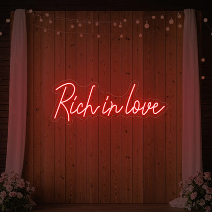 Photo of the 'Rich In Love' neon sign glowing in red against a styled interior wall.