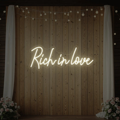 Photo of the 'Rich In Love' neon sign glowing in warm white against a styled interior wall.