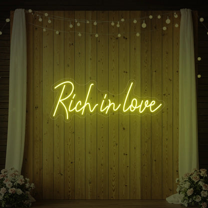 Photo of the 'Rich In Love' neon sign glowing in lemon yellow against a styled interior wall.
