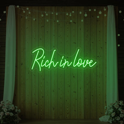 Photo of the 'Rich In Love' neon sign glowing in green against a styled interior wall.