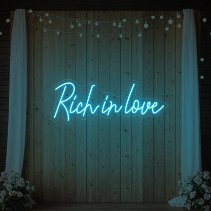 Photo of the 'Rich In Love' neon sign glowing in ice blue against a styled interior wall.