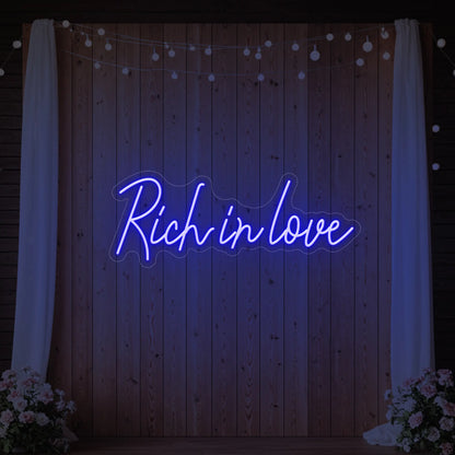 Photo of the 'Rich In Love' neon sign glowing in deep blue against a styled interior wall.