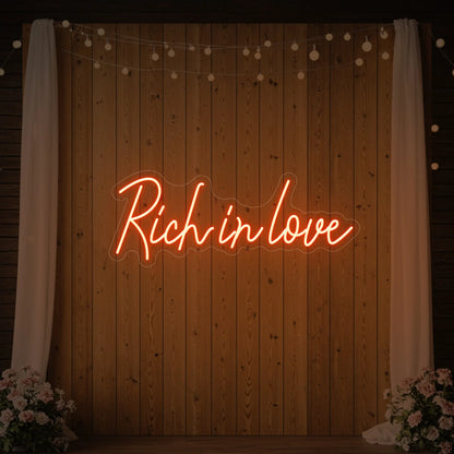 Photo of the 'Rich In Love' neon sign glowing in golden yellow against a styled interior wall.
