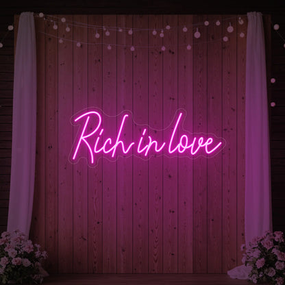 Photo of the 'Rich In Love' neon sign glowing in light pink against a styled interior wall.