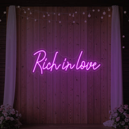 Photo of the 'Rich In Love' neon sign glowing in deep pink against a styled interior wall.