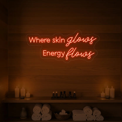 Photo of the 'Where Skin Glows, Energy Flows' neon sign glowing in golden yellow against a styled interior wall.