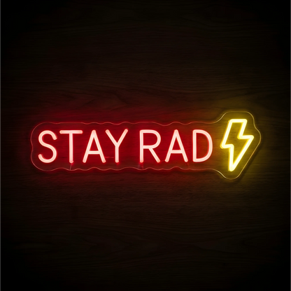 “STAY RAD” LED Neon Sign glowing in red uppercase letters with a yellow lightning bolt on clear acrylic against a dark wood background, suited to bedrooms and creative studios.