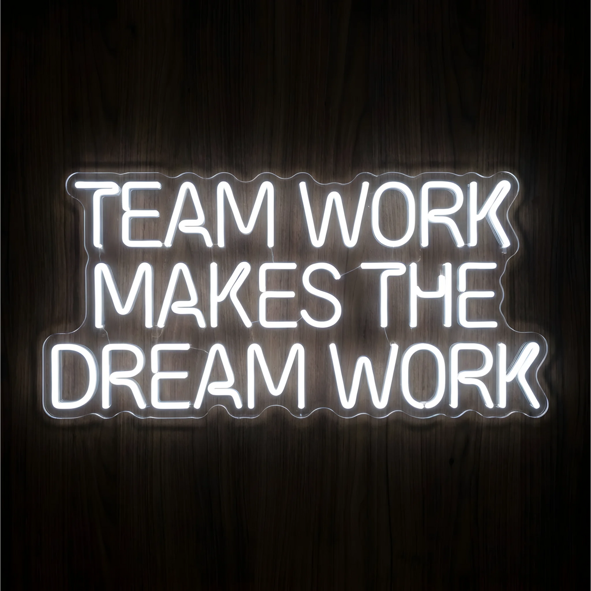 Teamwork Makes the Dream Work LED Neon Sign with bright white uppercase lettering across three lines on clear acrylic against a dark wood background, ideal for offices and collaborative workspaces.