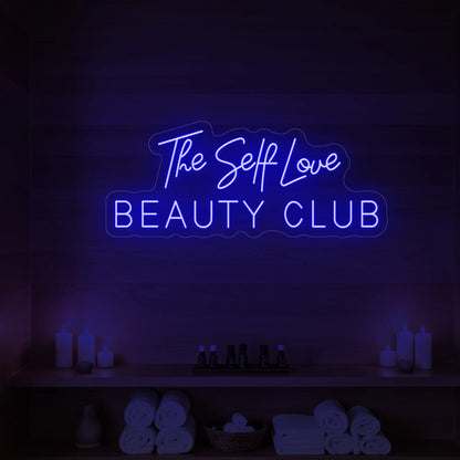 Photo of the 'The Self Love Beauty Club' neon sign glowing in green against a styled interior wall.