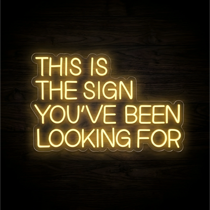 This Is the Sign You've Been Looking For LED Neon Sign displaying “THIS IS THE SIGN YOU’VE BEEN LOOKING FOR” in warm yellow uppercase lettering on contoured clear acrylic backing against a dark wood background, suited to living rooms and home offices.