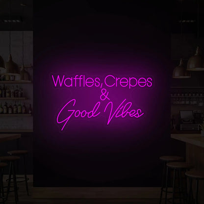 Photo of the 'WAFFLES, CREPES & GOOD VIBES' neon sign glowing in deep pink against a styled interior wall.