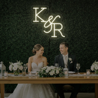 Photo of the 'Wedding Initials Custom Neon Sign V2' neon sign glowing in warm white against a styled interior wall.