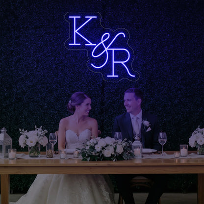 Photo of the 'Wedding Initials Custom Neon Sign V2' neon sign glowing in deep blue against a styled interior wall.