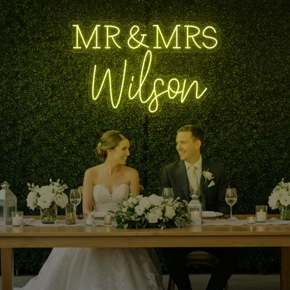 Photo of the 'Wedding Last Names Custom Neon Sign V2' neon sign glowing in lemon yellow against a styled interior wall.