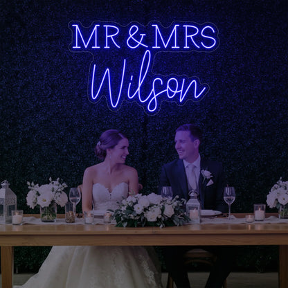 Photo of the 'Wedding Last Names Custom Neon Sign V2' neon sign glowing in deep blue against a styled interior wall.