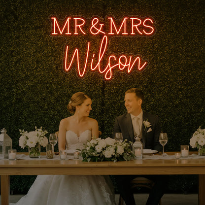Photo of the 'Wedding Last Names Custom Neon Sign V2' neon sign glowing in golden yellow against a styled interior wall.