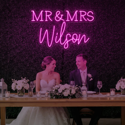 Photo of the 'Wedding Last Names Custom Neon Sign V2' neon sign glowing in light pink against a styled interior wall.