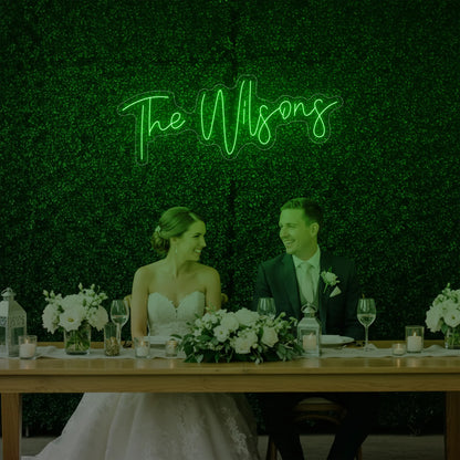 Photo of the 'Wedding Last Names Custom Neon Sign V4' neon sign glowing in green against a styled interior wall.