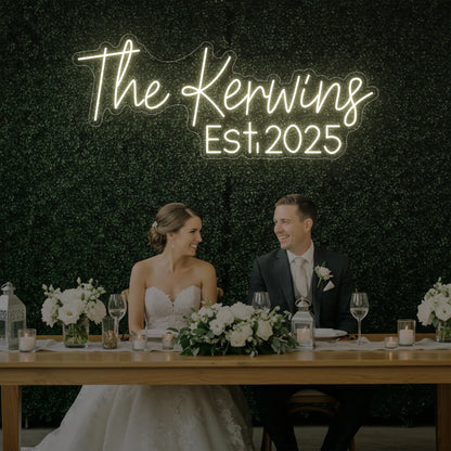 Photo of the 'Wedding Last Names Custom Neon Sign V5' neon sign glowing in warm white against a styled interior wall.
