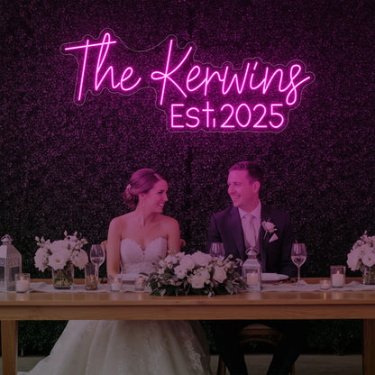 Photo of the 'Wedding Last Names Custom Neon Sign V5' neon sign glowing in light pink against a styled interior wall.