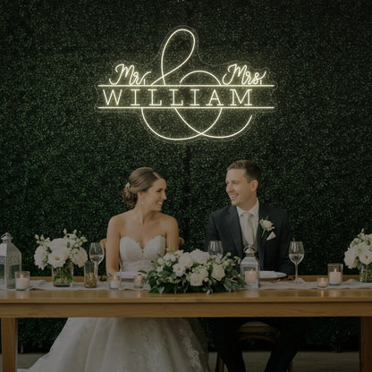 Photo of the 'Wedding Last Names Custom Neon Sign V3' neon sign glowing in warm white against a styled interior wall.