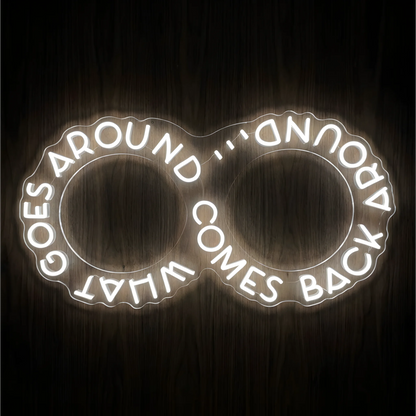 What Goes Around Comes Back Around LED Neon Sign in glowing white lettering arranged around two outlined circles on clear acrylic against a dark wood background, suited to home offices and living rooms.