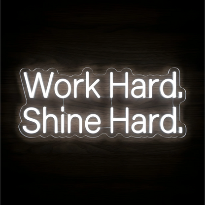 Work Hard Shine Hard LED Neon Sign with bright white lettering on clear acrylic against a dark wood background, suited to home offices and motivational workspaces.