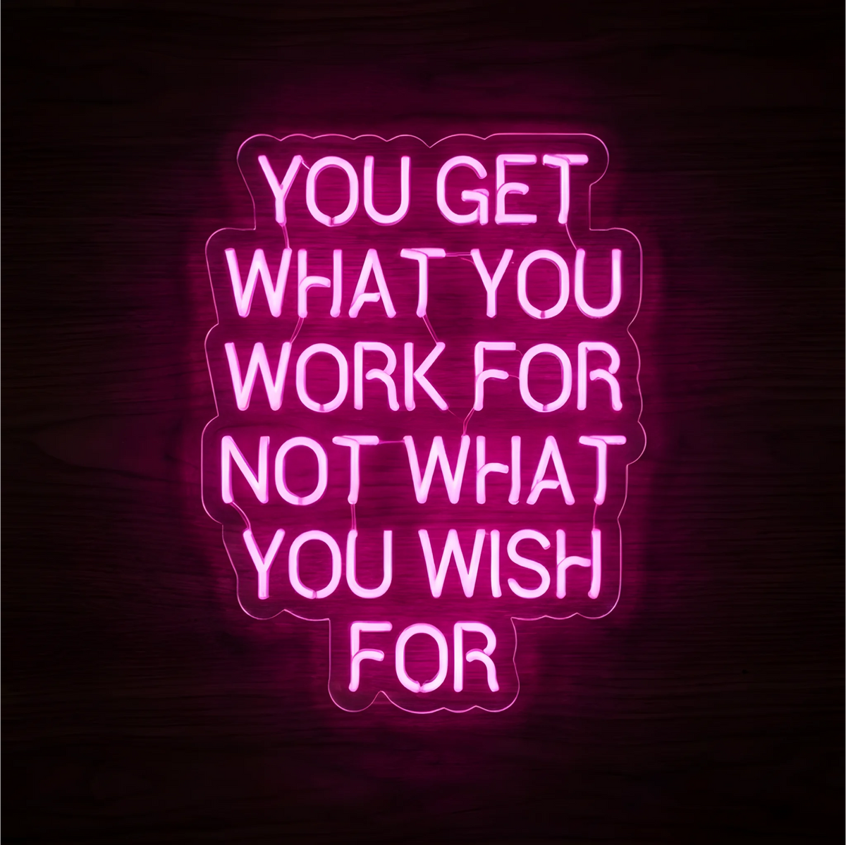 “You Get What You Work For Not What You Wish For” LED Neon Sign glowing in bright pink on clear acrylic backing against a dark wood background, suited to home offices, workspaces, and motivational walls.