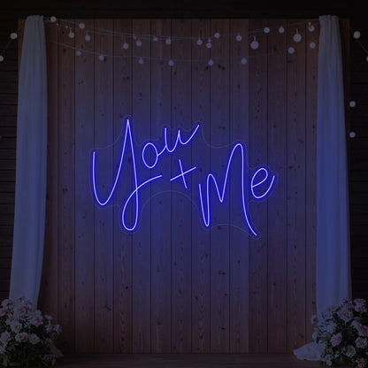 'You + Me' neon sign glowing in deep blue against a styled interior wall.